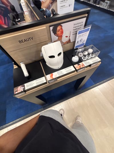 A retail display table, shot from an elevated angle, showcases beauty products. The word "BEAUTY" is vertically printed on a wood-grained surface. A white, futuristic-looking helmet rests on the table. Accompanying displays feature promotional materials with images of people using the products. A person stands nearby, visible from the knees down, wearing grey sweatpants and sneakers. The table sits on a blue carpet next to a lighter-colored flooring.