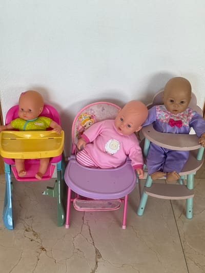 Three baby dolls are seated in three individual toy high chairs, arranged in a line from left to right, against a plain white wall and on a light-colored, patterned tile floor. This scene is located in the city of Miramar, Mexico.
The leftmost doll has light skin, a bald head with a small pink bow, and blue eyes. It is dressed in a lime-green, short-sleeved top with white polka dots and the word "Nenuco" on the chest. This doll sits in a high chair featuring a pink plastic seat, a yellow plastic tray, and a base with light blue and green plastic components.
The central doll has light skin, a bald head, and blue eyes. It wears a long-sleeved pink top with a white sheep motif on the chest, paired with pink and white striped pants. This doll occupies a high chair with a pink metal frame, a purple plastic seat, and a purple plastic tray. The seat back is decorated with a pattern depicting a blonde princess.
The rightmost doll has a darker skin tone, a bald head, and dark eyes. It is clothed in a long-sleeved purple top with a multi-colored floral and heart pattern and a pink bow on the chest, along with purple pants. This doll is positioned in a high chair with a light blue and mint green frame, and light brown or beige seat and tray. Its bare feet are visible below the tray.
The floor beneath the high chairs consists of light-toned tiles with a distinct, dark, irregular vein pattern. The wall behind the objects is uniformly white with a slight textured finish.