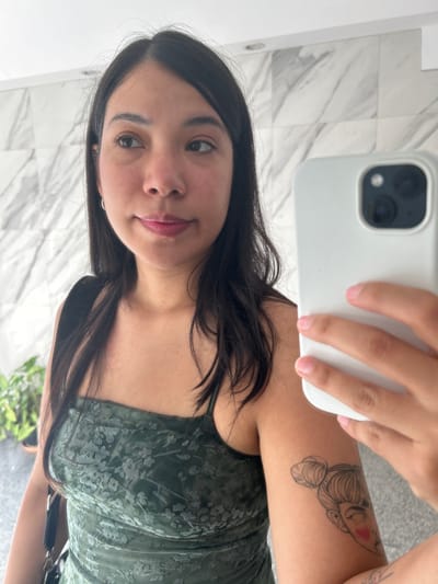 An image taken in Ciudad del Libertador General San Martín, Argentina, depicts a young adult female captured in a mirror selfie. The individual, of East Asian or mixed heritage, has long dark hair, dark eyes, and is wearing a dark green strapless or spaghetti-strap top or dress with a subtle floral or leaf pattern. A black strap from a bag is visible over her left shoulder. She has a small silver hoop earring in her left ear and light pink lipstick on her lips. Her right arm displays a black outline tattoo of a woman's head with two buns and a red cheek blush. She holds a white smartphone, likely an iPhone, in her right hand to take the photograph, with the phone's camera module facing the mirror. The background consists of light-colored, possibly white or grey, marble-patterned tiled walls. A potted green plant is partially visible in the lower-left corner of the frame.