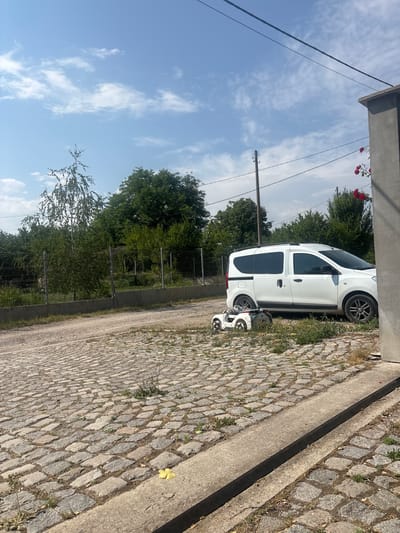 The image depicts a scene on a sunny day, likely in the city of Prolesha, Bulgaria. In the foreground, a cobblestone driveway leads towards a white van parked on the right side of the frame. In front of the van, two small toy cars are placed on the cobblestones, one white and one black. The background is filled with lush green trees and foliage, with a wire fence visible in the middle ground. The sky is a clear blue with scattered white clouds. A grey concrete pillar stands on the far right, with red roses growing on a vine next to it. The environment appears to be a residential or semi-rural area.