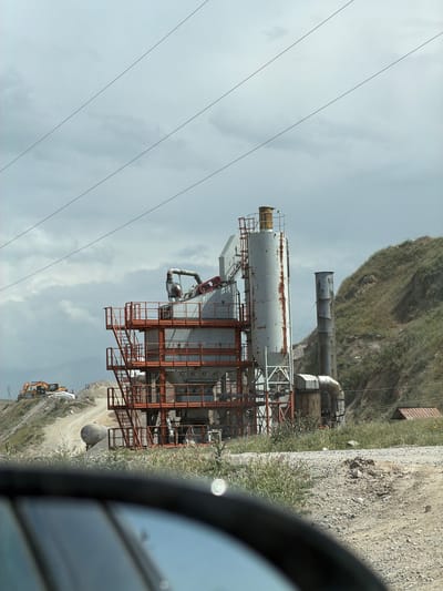 The image displays an industrial plant, likely involved in quarrying or asphalt/concrete production, situated in a mountainous landscape, consistent with the Баш-Кара-Суу region of Kyrgyzstan. The main subject is a large, weathered industrial complex featuring several tall, grey cylindrical tanks or silos, interconnected by extensive orange metal scaffolding, stairs, and various pipes. Rust is visible on the surfaces of the tanks. Behind this primary structure, a tall smokestack or chimney rises, with a steep, green-brown, grassy hillside forming the background.
The sky is overcast and cloudy, indicating daytime conditions. In the foreground, a dirt road or path runs alongside the plant. An excavator and possibly another piece of heavy construction machinery are visible parked on a dirt mound. A yellow "P" sign, indicating parking, is also visible near the machinery. Sparse vegetation grows along the roadside and on the hillside. Several power lines stretch across the top portion of the sky.
The photo appears to be taken from within a vehicle, with part of a car's rearview mirror obscuring the right side of the frame. There are no people visible in the image. The scene suggests an active or recently active industrial site in a rugged, natural setting. No discernible text other than the "P" sign is clearly visible.