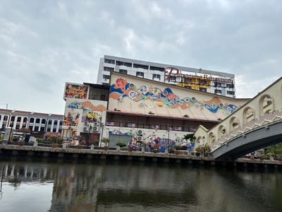 A waterside view displays a bridge spanning the canal. A building adorned with murals is visible along the water's edge. A taller structure with the inscription "Panglima Battlesawa" stands behind the mural-decorated building. Reflections ripple across the water's surface, mirroring the structures. The sky is overcast.