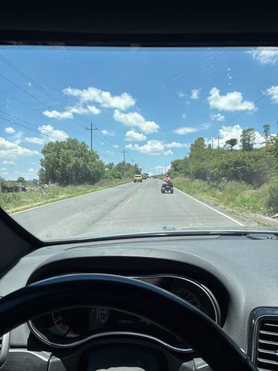This is a forward-facing, first-person view from inside a vehicle, likely a Jeep Grand Cherokee, looking through the windshield. The scene depicts a rural road in Santiago Tlautla, Mexico, on a clear, sunny day with scattered white clouds.
The road ahead is paved and appears to be a two-lane highway with a solid white line on the right edge and dashed yellow lines separating the lanes. The vehicle is in the right lane, suggesting it is traveling in the same direction as the vehicles ahead.
In the distance, on the road, are several vehicles. Closest is a four-wheeled ATV being ridden by a person wearing a pink shirt. Further ahead, there is a yellow vehicle, possibly a SUV or truck, and another smaller vehicle that is less discernible.
The environment surrounding the road is characterized by dry, grassy verges and dense green vegetation, including trees and bushes. Utility poles with power lines are visible along the side of the road, indicating the presence of infrastructure in the area. There are also some piles of debris and discarded materials on the left side of the road, suggesting the area may be less developed or maintained.
Inside the vehicle, the dashboard and steering wheel are visible in the foreground, with a partial view of the instrument cluster. The odometer shows "173921 km". The overall impression is of a typical drive through a semi-rural or suburban area in Mexico.