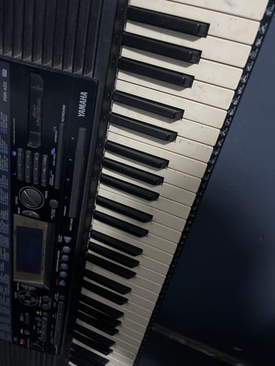 A Yamaha PSR-420 electronic keyboard is depicted at an angle in Alasia, Nigeria. The keyboard, primarily black, features cream-colored white keys and black black keys, exhibiting signs of wear and discoloration on the white keys. The control panel, located on the left side, displays the "YAMAHA" brand name, "PSR-420" model designation, a cartridge slot, a monochrome display screen, and numerous buttons and dials for functions such as Style, Voice, Tempo, and Volume. The keys extend from the lower-left to the upper-right of the frame. The background consists of a plain, dark blue or dark grey surface. No people or active interactions are visible in the scene.