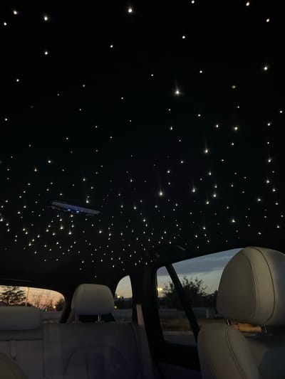 The image depicts the interior of a car, specifically looking upwards towards the ceiling and out of the rear windows. The car's ceiling is adorned with numerous small, illuminated lights that resemble stars. In the foreground, parts of light-colored leather car seats are visible. Through the rear windows, a dusky outdoor scene can be observed, with silhouetted trees and a muted sky, suggesting it is either dawn or dusk. There are no people visible in the image. The location is identified as Šempeter pri Gorici, Slovenia.