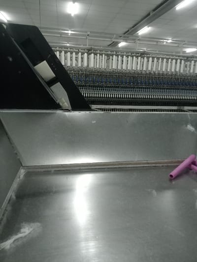 The scene depicts an interior view of an industrial facility, specifically a textile manufacturing area, located in Sukomoro, Indonesia. The foreground features a large, reflective, flat metal surface, possibly a collection tray or workbench, with a small amount of white fibrous debris visible on the lower-left edge. Two to three pink, tubular objects, resembling empty yarn tubes or bobbins, are situated on the metal surface in the lower-right quadrant. Directly beyond this surface, a section of black and gray industrial machinery is visible. The midground is dominated by a multi-tiered textile machine, displaying numerous rows of white spools or bobbins arranged horizontally, with visible thread or yarn extending downwards. The upper background shows the ceiling, equipped with multiple elongated fluorescent light fixtures, illuminating the workspace. No personnel or discernible human activity are present.