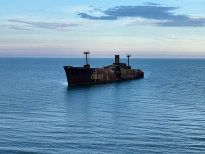 This forensic description details an outdoor scene depicting the shipwreck of the MV Evangelia located off the coast of Costinești, Romania. The dominant subject is a large, heavily rusted cargo ship, dark brown and black in color, positioned centrally in the calm, blue-green waters of the sea. The ship is stationary, partially submerged or beached, extending horizontally across the mid-ground of the frame. Its superstructure is severely degraded, with remnants of masts and deckhouses visible. Several seabirds are perched on the highest points of the ship's remaining structures. Faint, weathered lettering is discernible on the hull, including fragments like "DUCA" and "Summer Camp."
The ocean occupies the lower two-thirds of the image, displaying gentle ripples across its surface under a diffused light. The water's color transitions from a darker blue close to the foreground to a lighter, more aquamarine hue near the horizon. Above the distinct, straight horizon line, the sky is a blend of pale blue and light grey, featuring scattered, wispy white clouds and more substantial, darker grey-blue clouds, particularly in the upper right quadrant. The overall lighting suggests daylight, possibly late afternoon, with no direct harsh sunlight. No human figures are present in the scene.