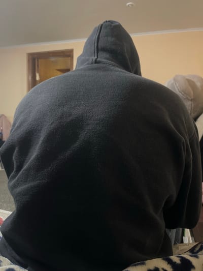 The image shows the back of a person wearing a black hoodie. The person is seated, and the hoodie appears to be made of a soft, possibly fleece material. The background includes a wall painted in a light, neutral color, a doorway with a wooden frame, and a glimpse of a ceiling light fixture. To the right, a blurred light-colored object, possibly a cushion or stuffed toy, is visible. The lighting suggests an indoor setting, likely a room in a residential building. The image is taken from behind the person, focusing on their upper back and head covered by the hoodie. There is no specific information to identify the location as Polonne, Ukraine.