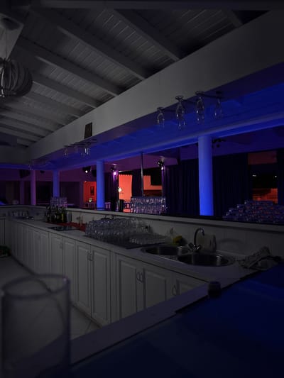 The image depicts the interior of a bar or restaurant with mood lighting. The ceiling is white and slatted, with a large, decorative light fixture visible on the left. Below the ceiling, a long white counter runs along the wall, equipped with a double sink and faucet. Numerous stacks of clean glassware, including wine glasses, are arranged on shelves above the counter and on the counter itself. Bottles of alcohol and a red cloth are also present on the counter. The background features a stage area with purple curtains, illuminated by red and blue lights. Columns support the structure, and the overall ambiance suggests a nightlife venue. The location is identified as Zverino, Bulgaria, though no specific landmarks within the city are discernible from the image. No people are visible in the scene.