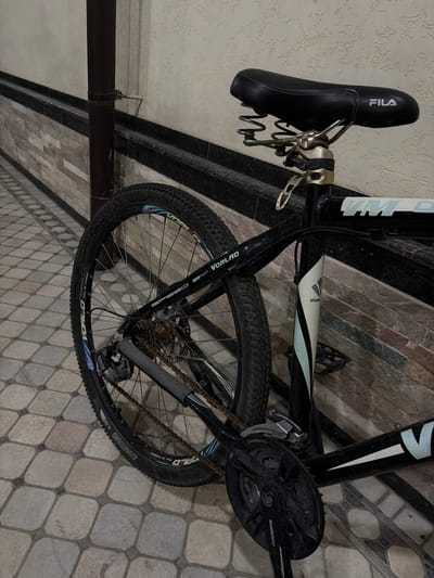 A black bicycle with a black Fila-branded seat is parked on a tiled patio. The bicycle has a black frame and black tires with blue and white accents. The rear wheel of the bicycle is visible, as is the chain and pedals. The bicycle is leaning against a textured wall with horizontal stone-like accents and a dark border. A dark, cylindrical pipe is to the left of the bicycle. The scene appears to be outdoors.