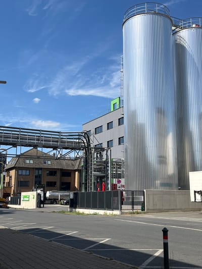 The image depicts an industrial scene, likely a factory or processing plant, under a clear blue sky with sparse white clouds. The main subjects are large, cylindrical silver storage tanks, typical of industrial facilities. One tank is prominently featured in the foreground, appearing corrugated and reflective. Behind it, a portion of another similar tank is visible.
A modern industrial building with a grey facade and multiple windows is situated adjacent to the tanks. A section of a brighter green structure is also visible on this building. Extensive pipework and infrastructure, including metal walkways and supports, are spread across the middle ground, connecting various parts of the facility.
In the background, a building with a darker, brick-like facade and more windows is partially visible. A tanker truck is parked in front of this building, suggesting ongoing operations or deliveries. A black bollard with red and white markings stands on an asphalt surface in the foreground, marking a parking area or boundary.
The scene appears to be taking place during the daytime, as indicated by the bright sunlight casting shadows. The weather is pleasant with a clear sky. The presence of industrial structures, the tanker truck, and the overall setting strongly suggest an industrial zone in Neuss, Germany. No discernible emotions are conveyed in the image; it is a factual representation of an industrial environment. There is some text visible on a green sign in front of the brick building, but it is too small and blurry to be readable. Another visible sign, red and white, near the bollard shows directional arrows, possibly indicating traffic flow or prohibition.