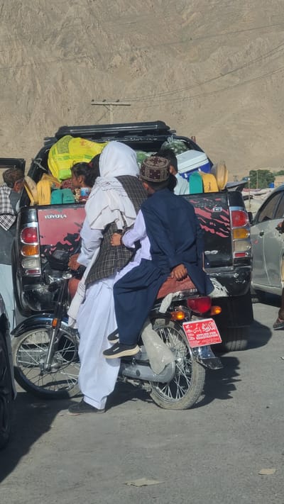 The scene depicts multiple individuals riding on a motorcycle, overloaded with luggage and provisions. The motorcycle has a license plate visible that reads "Applied For Registration" in both English and Urdu. The surroundings appear to be Bostan, Pakistan, given the arid environment and the style of dress of the people.