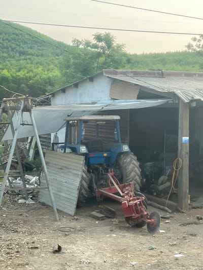 A blue tractor with a red attachment sits partially inside a shed structure. The shed has a corrugated metal roof and siding, with an open front. The surrounding environment is a dirt yard with scattered debris. In the background, a lush green hillside is visible. The photograph appears to have been taken near Myui, Vietnam.