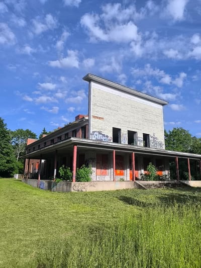The image depicts a long, low-lying abandoned building with a large covered porch stretching across its front. The building, constructed of brick, shows signs of disrepair and extensive graffiti on its walls and the porch. The porch itself is supported by red-painted pillars. Above the porch, the building has a second level with several open, dark window frames and is covered in horizontal siding. The roofline is somewhat unusual, with a taller section at the front.

The surrounding area is overgrown with lush green grass and foliage, with mature trees visible on the left side of the building and in the background. The sky is a vibrant blue, dotted with scattered white clouds, suggesting a pleasant, possibly midday, weather condition.

There is no activity or event taking place; the scene conveys a sense of stillness and abandonment. Notable details include the graffiti, the dilapidated state of the building, and the contrast between the neglected structure and the lively natural surroundings. The location context is Rannamõisa, Estonia. No legible text is clearly discernible within the image.