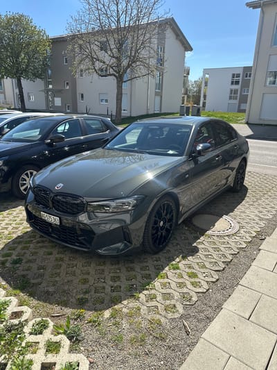 The image shows a gray BMW parked on interlocking concrete pavers in a residential area of Jestetten, Germany. A black vehicle is partially visible to the left of the BMW. Buildings and trees are in the background under a blue sky. The license plate of the BMW reads "AG-575363". A manhole cover is near the front tire of the BMW.