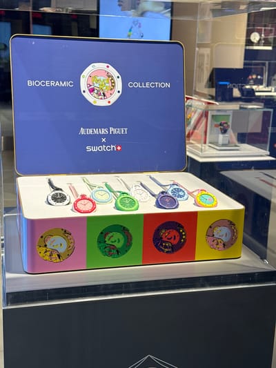 The image shows a display case containing two boxes of Audemars Piguet x Swatch Bioceramic MoonSwatch watches. The box on the left is dark blue and features the Audemars Piguet logo. The box on the right is white with colorful circular designs, each corresponding to a different model of the MoonSwatch. The display case appears to be located in Oak Brook, United States of America, based on the presence of the Audemars Piguet x Swatch collaboration which are sold in authorized boutiques and pop up stores. No people are visible.