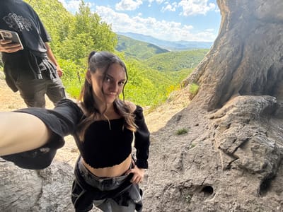 A low-angle, rotated outdoor photograph captures a woman, seemingly taking a selfie, with a scenic backdrop. The woman has dark hair and is wearing a black crop top with long sleeves. She is positioned in the upper-middle of the frame, with a small smile.

To the left, there's a rocky overhang with visible texture and erosion patterns. In the background, a lush green valley stretches out, dotted with trees and vegetation. In the distance, a vista of rolling hills and a partly cloudy sky are visible. Another person, with a black top, is also visible.

The location is likely in Lipnitsa, Bulgaria.