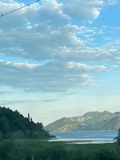 The image depicts a landscape with a cloudy sky dominating the left side. To the right, a body of water is bordered by mountainous terrain and a dense treeline. This scene is likely in Braćeni, Montenegro.