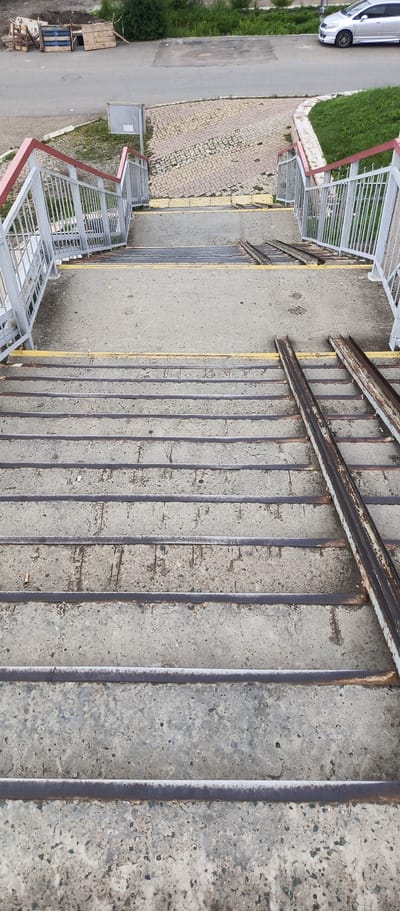 The image shows a downward view of a concrete pedestrian overpass in Pogranichny, Russia. The overpass features metal railings with red tops and yellow warning strips on the stairs. Two sets of rusty metal ramps are installed alongside the concrete stairs. The environment includes a paved road and some greenery visible at the top of the frame, with a silver car parked on the side of the road. The concrete stairs are worn and show signs of weathering and some debris.