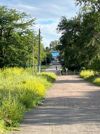 A paved path winds through a park with abundant green trees and lush vegetation, including a vibrant patch of yellow wildflowers on the left. The path is sparsely populated with people: a person on a scooter is visible in the mid-ground, and further up, another person is walking with a stroller, and another individual is walking a dog. In the background, a modern, light blue building with a white facade stands at the end of the path, possibly a community center or public facility. The sky above is a mix of blue with scattered white clouds, indicating a pleasant day. The scene appears to be a well-maintained urban park, likely in Shyroka Dacha, Ukraine.