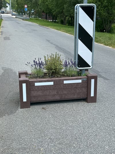 The image shows a street scene in Le Brassus, Switzerland, with a focus on a brown planter box containing lavender and a bush. A tall sign with black and white diagonal stripes is positioned behind the planter. The street is paved and appears to be wet in some areas, indicating recent rain. In the background, trees and buildings are visible, along with other road signs. A few cars and a yellow bus are parked or driving on the road in the distance. The overall scene depicts an outdoor, urban environment in a mountainous region, likely during daytime given the lighting conditions.