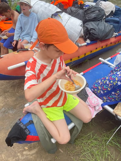 The image shows a group of children on an inflatable raft on a muddy riverbank. The central subject is a young boy wearing an orange cap and a red and white striped t-shirt, eating pasta from a bowl. To his left, another boy, wearing a white hat and a blue hooded shirt, is also holding a bowl. Several large bags, some black and some silver, are piled on the raft behind the children. The riverbank is covered in mud and sparse green grass. The overall setting appears to be outdoors, likely during a warm season. There is no indication of the location being in the city of Нежинка, Russia.