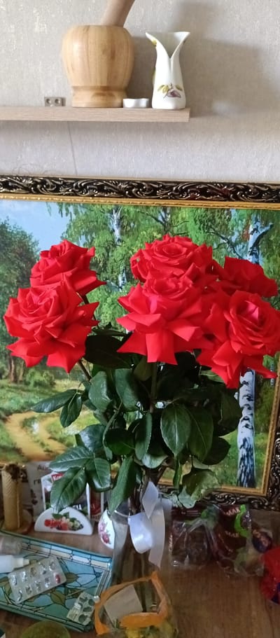 A still-life photograph taken indoors. Dominating the scene is a bouquet of bright red roses with green leaves in a clear glass vase tied with a white ribbon. Behind the flowers is a framed painting depicting a forest landscape. On a shelf above the painting are a wooden mortar, a white ceramic vase, and a small white candle. Beneath the roses, on a cluttered surface, are various objects including medicine packaging and other unidentified items. The photograph provides no evidence to confirm or deny it was taken in Irbit, Russia. No people are visible.