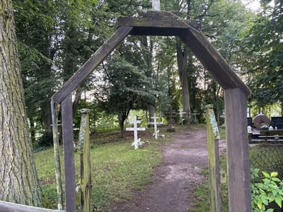 This image depicts the entrance to an Orthodox cemetery in Wojnowo, Poland. The foreground is dominated by a weathered, dark wooden archway forming a gateway, with a large tree trunk visible on the left, displaying textured bark with green lichen. A narrow, unpaved dirt path leads from the archway into the cemetery.
In the midground, numerous grave markers are visible, predominantly Orthodox crosses characterized by their distinctive three-barred design. Several white crosses stand upright on the left side of the path, while darker, more weathered crosses are dispersed further back. Green grass covers the ground between the graves. On the right, beyond the path, a distinct circular stone object resembling a millstone is present on top of a grave, with other headstones and a partially visible chain-link fence nearby.
The background consists of dense deciduous trees and foliage, indicating a wooded or heavily vegetated environment surrounding the cemetery. The scene is illuminated by natural light, suggesting daytime conditions. No individuals or active interactions are present within the frame.