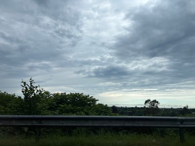 The image depicts a cloudy sky over a landscape in Kveda Kvirike, Georgia. In the foreground, a metal guardrail runs horizontally across the frame, with green grass and foliage beneath it. Beyond the guardrail, lush green trees and vegetation dominate the middle ground. In the distance, the horizon shows a scattering of trees, some buildings, and a faint line that could be water or a distant coastline. The sky is filled with heavy, gray clouds, suggesting an overcast or impending rainy day. There are no people or animals visible in the scene.