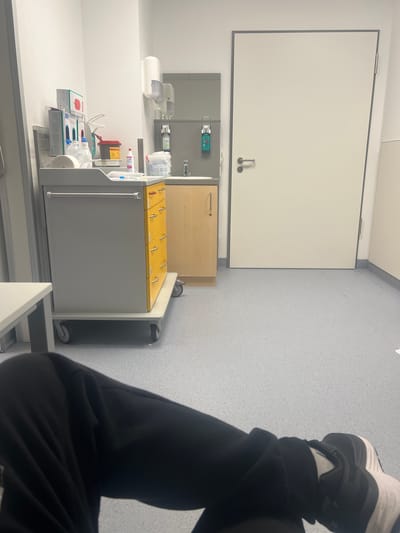 The image captures an indoor scene, likely within a medical facility in Herne, Germany. The perspective is from a seated or reclining person, showing their legs and feet in the foreground on the right, wearing black sweatpants and white and black athletic sneakers.
The main focus of the background is a functional, clean environment. To the left, a large, rectangular white cabinet or panel with a grey trim is mounted on the wall, featuring two handles in the lower right portion. Below this, there's a counter area with a light wood-colored cabinet and a grey unit. On the grey unit, a green wall-mounted hand sanitizer dispenser is visible. Further left on the counter, there's a white tissue dispenser and various medical supplies.
A prominent grey medical cart or trolley with yellow-lidded compartments is positioned in the center-right. Several of these yellow lids have small handles, and at least one is marked with the number "1". The cart is equipped with various medical items, including packaged intravenous cannulas (with visible text "Vasofix® Safety IV cannula" and "B.BRAUN"), disinfectants, a red sharps container, and other supplies commonly found in a treatment room.
The floor is a light grey with a speckled texture, and the walls are a plain light color, with some sections featuring a subtle textured wallpaper. The lighting suggests an interior during daytime, providing clear visibility of all objects. No specific activity is actively occurring beyond the presence of the individual, who appears to be resting or waiting. The overall impression is clinical, organized, and sterile.
