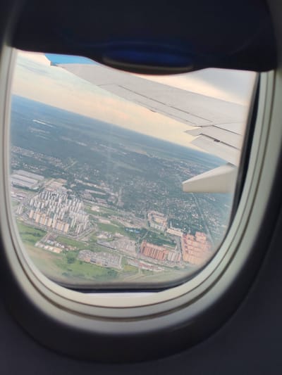 This image, taken from an airplane window, shows an aerial view of a city, specifically the area around Khrabrovo, Russia. A portion of an airplane wing is visible in the upper right of the frame. Below the wing, the city unfolds with a mix of residential buildings, industrial structures, and green spaces. Dominating the central part of the image are several clusters of multi-story apartment buildings, some with light-colored facades and others with orange or brown brickwork. Surrounding these residential areas are smaller buildings, roads, and patches of trees and grass. In the background, the urban sprawl extends towards the horizon, meeting a pale sky. The overall impression is of a suburban or semi-urban landscape viewed from a significant altitude.