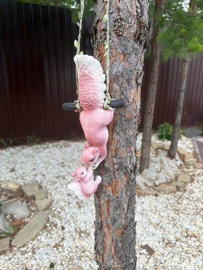 Two pink resin squirrel figurines are hanging upside down from a string wrapped around a pine tree in what appears to be a backyard or garden setting. The larger squirrel, positioned above, has its tail raised and curved over its head, appearing fluffy and textured with white and pink. It is grasping a dark horizontal rod with its paws. Below the larger squirrel, a smaller squirrel is also hanging, appearing to be interacting with the larger one, possibly reaching up towards its face. This smaller squirrel also has pink coloring and textured white accents on its tail and ears.
The pine tree has rough, textured bark, and the string holding the figurines is adorned with small green leaf-like embellishments. In the background, a dark brown wooden fence runs horizontally across the image. Behind the fence, there are visible green pine branches and what appears to be a darker, blurred area. The ground in the foreground is covered with a layer of small white pebbles and scattered, light-colored rocks of varying sizes. Several other pine trees are visible in the mid-ground, with the one in the foreground being the most prominent. The overall scene suggests an outdoor decorative arrangement.