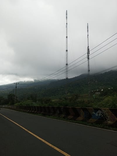 The scene depicts an overcast day in the city of Deothang, Bhutan, with a paved road in the foreground curving slightly right. A solid yellow line marks the visible road segment. To the right of the road, a segmented concrete barrier, featuring dark bases and faded lighter chevron patterns, is present, with unreadable graffiti on some segments. Beyond the barrier, dense green vegetation borders the road. A dark utility pole stands on the left, supporting multiple overhead power lines that traverse the midground. Two tall, slender, metallic lattice communication towers, equipped with various modules and supported by guy-wires, rise from the distant, heavily forested mountainside. The mountains are shrouded in mist or low-hanging clouds, blending with the uniformly grey sky. A faint, indistinct sign or structure is visible on the hillside beneath the right tower. No individuals are present.
