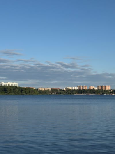 The image depicts a wide body of water, likely a river or lake, with a cityscape in the background. The water's surface is rippled and reflects the sky. Beyond the water, a dense line of green trees forms a natural barrier between the water and the urban area. A row of multi-story buildings, varying in architectural style and color, is visible along the treeline. Some buildings are white and modern, while others have red and orange facades, suggesting a mix of residential and possibly commercial structures. The sky above is a clear, pale blue with scattered, wispy white clouds. The overall scene suggests a tranquil environment where nature and urban development coexist. The location is identified as Osipovo, Russia. There are no visible people or specific actions taking place.
