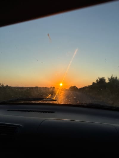 The image captures a sunset viewed from inside a vehicle, likely on a road trip or commuting. The sun, a bright orb of orange, is low on the horizon, casting a warm glow across the sky. The sky transitions from a pale blue at the top to a soft yellow and orange as it nears the horizon, indicative of the golden hour. Silhouetted against the sunset are trees and foliage, suggesting a rural or semi-rural setting.

The foreground is dominated by the interior of the car. The dashboard is visible on the right side of the image, appearing dark and textured. A windshield wiper is also visible, suggesting the car is in motion or has recently been used. The windshield itself has some smudges and streaks, typical of daily use, and a small, reddish smudge is noticeable on the left side of the glass. A bright streak of light refracts from the sun, extending upwards, likely due to dust particles or imperfections on the windshield.

The overall mood of the image is serene and tranquil, evoking the peacefulness often associated with sunsets. The colors are warm and inviting. The location context provided, Tankovo, Bulgaria, places this scene in a specific geographical area. No visible text is present in the image.