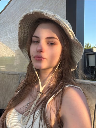 A young woman with long, light brown hair, wearing a white bucket hat and a white tank top with a cross necklace, is pictured in a close-up shot. The sun casts a shadow across her face, illuminating her lips and cheek. She is seated on a textured sofa outdoors, with a shaded awning and a bright blue sky visible in the background, suggesting a daytime setting. The environment appears to be a residential outdoor area.