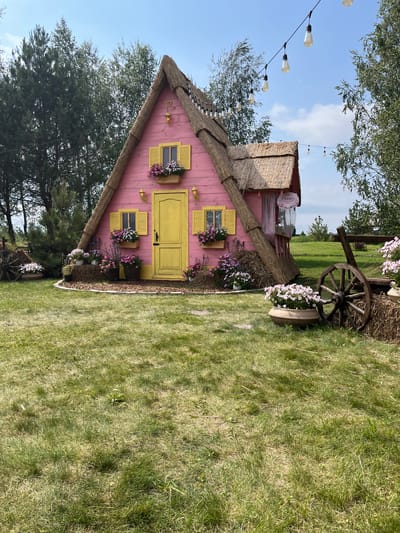 The image depicts a whimsical, pink, A-frame house with a thatched roof, situated in an outdoor setting. The house features yellow shutters on its windows and a bright yellow door. Flower boxes filled with pink and white flowers adorn the windowsills. String lights are suspended above the house, casting a festive ambiance. The house is surrounded by a grassy lawn, with trees visible in the background. To the right of the house, an old wooden wagon wheel is prominently displayed next to a bale of hay and a large planter filled with pink flowers. To the left, another wagon wheel is partially visible near a bale of hay with flowers. A small sign is attached to the house near the right side, reading "Peony". The scene suggests a charming, possibly decorative, outdoor space, consistent with a garden or event venue. The location is identified as Severynivka, Ukraine. There are no people visible in the image.