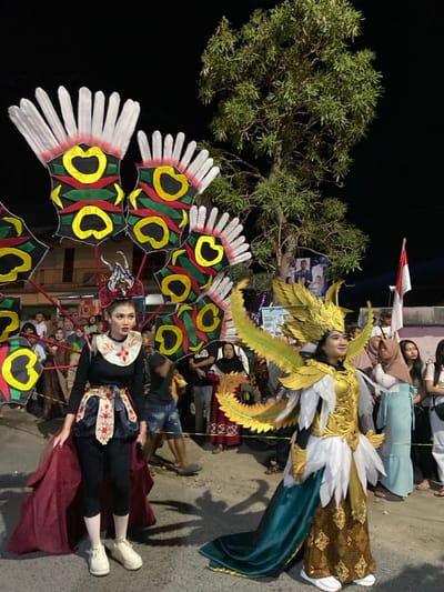 This image depicts a nighttime cultural event or parade taking place in the city of Kedungbanteng, Indonesia. The central focus is two individuals adorned in elaborate traditional or fantasy-inspired costumes, standing on a paved street.

The person on the left is a young woman wearing a black long-sleeved top, black leggings, and white sneakers. Over her top, she has a white, embroidered decorative collar and a multi-layered skirt with red, white, and black elements, featuring a long, flowing dark red train. Her headpiece is intricate, metallic, and predominantly red and black. She stands facing slightly right, with a direct gaze. Behind her, a large, fan-like structure composed of multiple segments, each featuring red, green, and yellow heart-shaped or oval designs with white feathered tops, is visible. This structure appears to be carried or is part of a larger float.

The person on the right is also a young woman, wearing a gold and white costume that resembles an angelic or mythical bird figure. This includes golden breastplate armor, white feathery elements on her torso and shoulders, and large, ornate green and gold wings extending from her back. Her lower garment consists of a brown patterned skirt with a long, flowing dark teal or green train. She wears white sneakers and a golden winged headpiece. She stands facing slightly left, smiling gently and looking towards the left.

In the background, a crowd of onlookers is visible, observing the procession. The environment is dark, indicating night, with artificial lighting illuminating the scene. A large tree with dense foliage is prominent in the upper right. Further back, buildings and what appears to be an Indonesian flag on a pole are discernible, along with some political campaign banners featuring faces, indicating a public street setting. A yellow rope or tape runs across the mid-ground, possibly sectioning off the parade route.