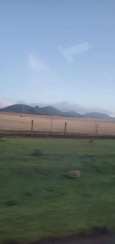The image depicts a rural landscape in what is presumed to be Kisima Ward, Kenya, though this cannot be confirmed from visual elements alone. The scene consists of a clear blue sky with some wispy cloud formations at the top of the image. Adjacent to the sky, there are mountains and in front of those is a field, possibly of dry grass or cultivated land, with a wooden fence running along the edge. To the right is a patch of green grass with a single rock. The camera angle is slightly tilted, giving the view the impression of being slightly off balance. Due to the resolution and potential camera movement, details are somewhat blurred.