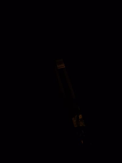 The image, taken in Le Teich, France, depicts a dimly lit scene focused on a metallic object. The object appears to be a pair of tongs or tweezers, made of a dark metal, possibly steel or iron. It is angled diagonally across the frame, with its jaws open. One jaw is slightly blurred, suggesting movement. The other jaw is more in focus and shows a textured surface, possibly with some discoloration or residue. The background is predominantly black, indicating a dark or underexposed environment, with faint, warm light reflecting off the metallic surface, highlighting its shape and texture. The overall impression is one of a close-up shot of an industrial or tool-like object in a very low-light setting.