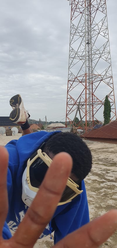 A person wearing a blue hoodie and protective goggles is shown from the chest up, with their hands in the foreground, obscuring their face. Their right leg is extended upwards, with a white and black sneaker visible. In the background, a large, red and white lattice tower stands tall against an overcast sky. The scene appears to be on a rooftop in Anguldi, Nigeria, with various buildings and a tall green tree visible in the distance. The ground is a flat, textured surface, possibly concrete or asphalt.