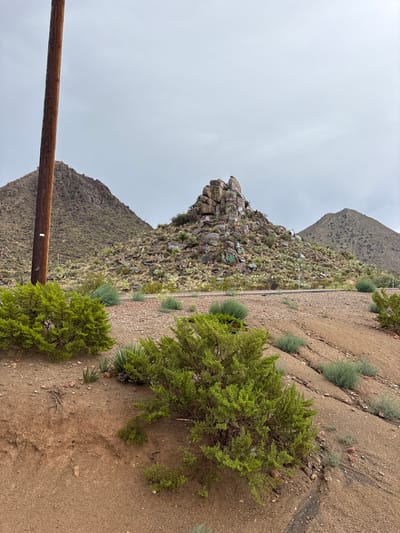 The image depicts a rocky, arid landscape with sparse vegetation, characteristic of a desert environment. In the foreground, a large wooden utility pole stands prominently to the left of the frame. The terrain slopes downwards and to the right, with patches of green shrubs and dry, sandy soil.

In the midground, a road or track cuts horizontally across the scene, bordered by low-lying plants and scattered rocks. Further back, a prominent rocky hill is covered in a mix of rocks and desert flora, including cacti. Several boulders on this hill are marked with what appear to be graffiti or colorful spray paint. Behind this hill, two larger, barren mountains rise in the background under an overcast sky. The overall impression is one of a remote, undeveloped area. The image does not contain any people or evidence of recent human activity beyond the graffiti on the rocks and the utility pole. The location is identified as being in the city of Etholen, United States.