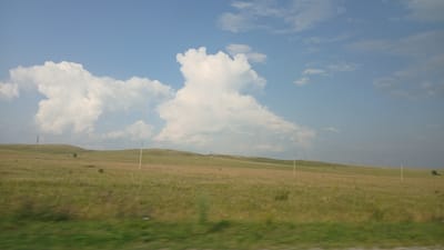 The image depicts a wide, rolling grassy plain under a bright blue sky with scattered cumulus clouds. Several utility poles are visible in the distance, spaced across the landscape, suggesting the presence of infrastructure. The foreground shows a blur of green grass, indicating movement, possibly from a vehicle. The overall scene suggests an open, rural environment, consistent with the steppe-like landscapes found in regions such as Krasnoshchyokovo, Russia. No people or animals are visible in the frame.