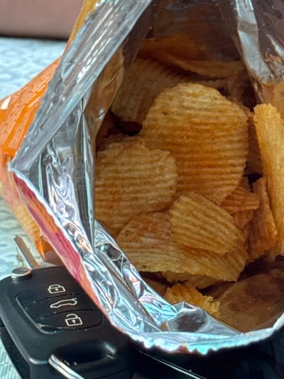 This is a close-up, indoor, eye-level shot of a partially opened bag of ridged potato chips. The bag is metallic and orange, with the chips spilling out. A black car key fob with three buttons is visible in the lower-left corner of the frame. The background is out of focus and appears to be a textured surface, possibly a tablecloth. There are no people or animals visible in the image. The scene is consistent with an outdoor setting on a warm day, given the apparent sunlight and casual placement of the items. The car key fob suggests the owner may have been driving or is about to drive. The context of Pribrezhnyi, Russia, indicates a potentially coastal environment, though the image itself does not provide any direct visual cues to confirm this. The overall impression is one of a brief, spontaneous pause, possibly a snack break during a journey.