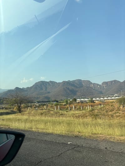 This image captures a wide-angle landscape view in Oacalco, Mexico, taken from inside a vehicle. The foreground features a dark asphalt road surface on the right, exhibiting minor cracks. On the lower left, the black frame and a portion of the red body of a vehicle's side mirror are visible. The midground consists of an expansive field predominantly covered in dry, straw-colored grass with scattered greener vegetation. A series of approximately ten light-colored, vertical posts, likely forming a fence line, extends horizontally across the field. To the left, a single leafless, deciduous tree stands. Beyond the field, a small cluster of buildings is discernible, including a dark-roofed structure and a prominent row of about ten light-colored, possibly residential or resort-style, structures characterized by rounded or conical roofs. The background is dominated by a substantial range of rugged mountains, appearing arid and covered with sparse, dark vegetation, stretching across the horizon. The sky above is clear and bright blue, with a few faint white clouds present towards the left. Reflections from a vehicle's windshield are visible across the upper portion of the image. No individuals or explicit human actions are depicted within the scene.