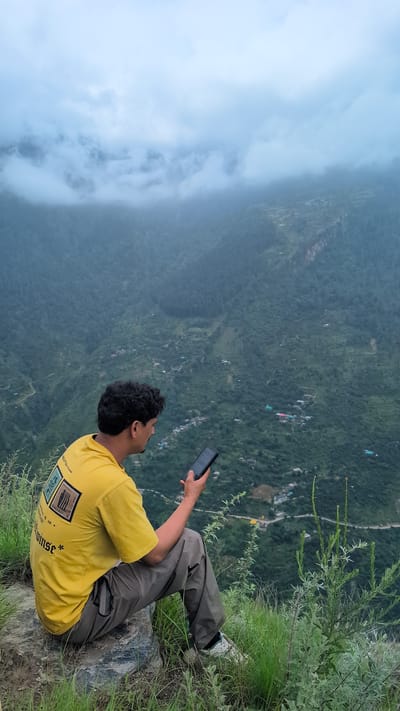 A man with dark, curly hair is sitting on a rock on a grassy cliff edge, looking at his smartphone. He is wearing a yellow t-shirt and grey pants. In the background, a vast, green, mountainous landscape is partially obscured by clouds. A winding road can be seen in the distance, along with scattered buildings that form a small village. The image appears to be taken in a rural, mountainous area, likely in or near Cheelaage, India.