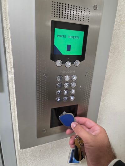 A close-up, eye-level shot captures a hand holding a blue key fob and keys, presented to a silver intercom system. The intercom, with a keypad and a green screen displaying "PORTE OUVERTE" (meaning "door open" in French), is mounted on a textured wall in what appears to be an urban building. The environment suggests a residential or commercial building entrance, possibly in Montrouge, France, given the presence of French text on the screen. The hand, with visible hair on the forearm, is poised to insert the key fob into the designated reader slot on the intercom. The scene is illuminated by ambient light, casting subtle shadows and highlighting the metallic sheen of the intercom and the worn texture of the wall.