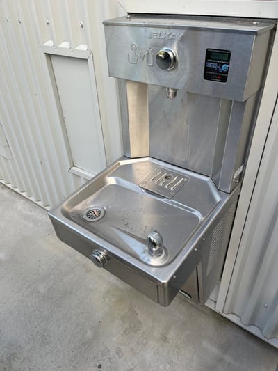An outdoor stainless steel water fountain and bottle filler station is the primary subject of this image, captured in Kelvin Grove, Australia. The unit is composed of two main sections: a bottle filler on the left and a traditional drinking fountain on the right.
The bottle filler section, branded "ELKAY EZ H2O", features a digital display showing a count of "1808625" with the accompanying text "DISPOSABLE PLASTIC BOTTLES SAVED", highlighting its environmental contribution. Below the display, a circular chrome-plated button activates the bottle filling spout, and a bottle-shaped outline is etched into the steel surface to guide users. A separate, lower spout for drinking is visible on the far left, complete with a push-button activator.
The right side of the unit contains a conventional drinking fountain, identified by its angled spout and a curved basin with a metal drain cover. Small water droplets are visible on the basin's surface, suggesting recent use. This fountain also has a chrome-plated push-button for activation.
The entire station is mounted against a corrugated white metal wall, indicative of a building exterior. The ground beneath the unit is a flat, grey concrete surface. The scene is brightly lit by natural daylight, suggesting a clear day. No people or activities are present, showing the facility in a static state. The station appears clean and well-maintained.