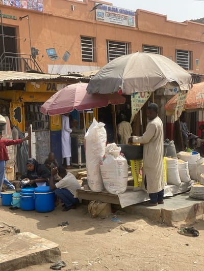 The image presents an active outdoor market scene in Rijiyar Lemo, Nigeria, under bright daylight. In the background, a two-story orange building stands with a sign for "DEFAH GLOBAL ENTERPRISES," indicating services like P.O.S, ATM, computers, CCTV, and mobile accessories. Painted murals of a laptop, a tablet, and a power plug are visible on the building's facade, along with a partial "SOMATU PLAZA" sign.
The mid-ground features several market stalls with corrugated metal roofs, shaded by large market umbrellas, including red/maroon and light grey/beige ones. On the right, a man in a long, light-beige traditional garment stands on a raised concrete platform, handling what appears to be grain from a large dark basin. He is positioned among multiple large white sacks, some labeled "U.A. Follia," and other beige sacks, indicating the sale of agricultural produce like rice and corn.
To the left, a yellow-walled stall displays mobile phone accessories, with a person in a white traditional garment visible inside. In the foreground, a woman in a dark head covering is seated on the ground, attending to three blue plastic coolers or buckets and a white bowl. Next to her, a man in a light t-shirt and dark pants crouches. A child in a red shirt and blue pants stands nearby, gesturing with an outstretched arm. The ground consists primarily of uneven, dusty earth with concrete sections and scattered debris. The scene depicts a bustling commercial area with various vendors and public activity.