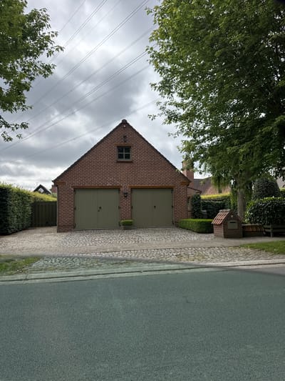 A two-door brick garage with light green doors sits in front of a residential property. The garage has a red brick facade, a gabled roof with dark shingles, and a small square window on the upper front wall. The driveway leading to the garage is paved with cobblestones. To the left of the garage, a tall green hedge forms a boundary. To the right, there is a small brick mailbox, a bench, and more manicured greenery. In the background, other houses and trees are visible, under a cloudy sky. Electrical wires are strung across the upper portion of the image. This scene is located on a street in Deerlijk, Belgium.